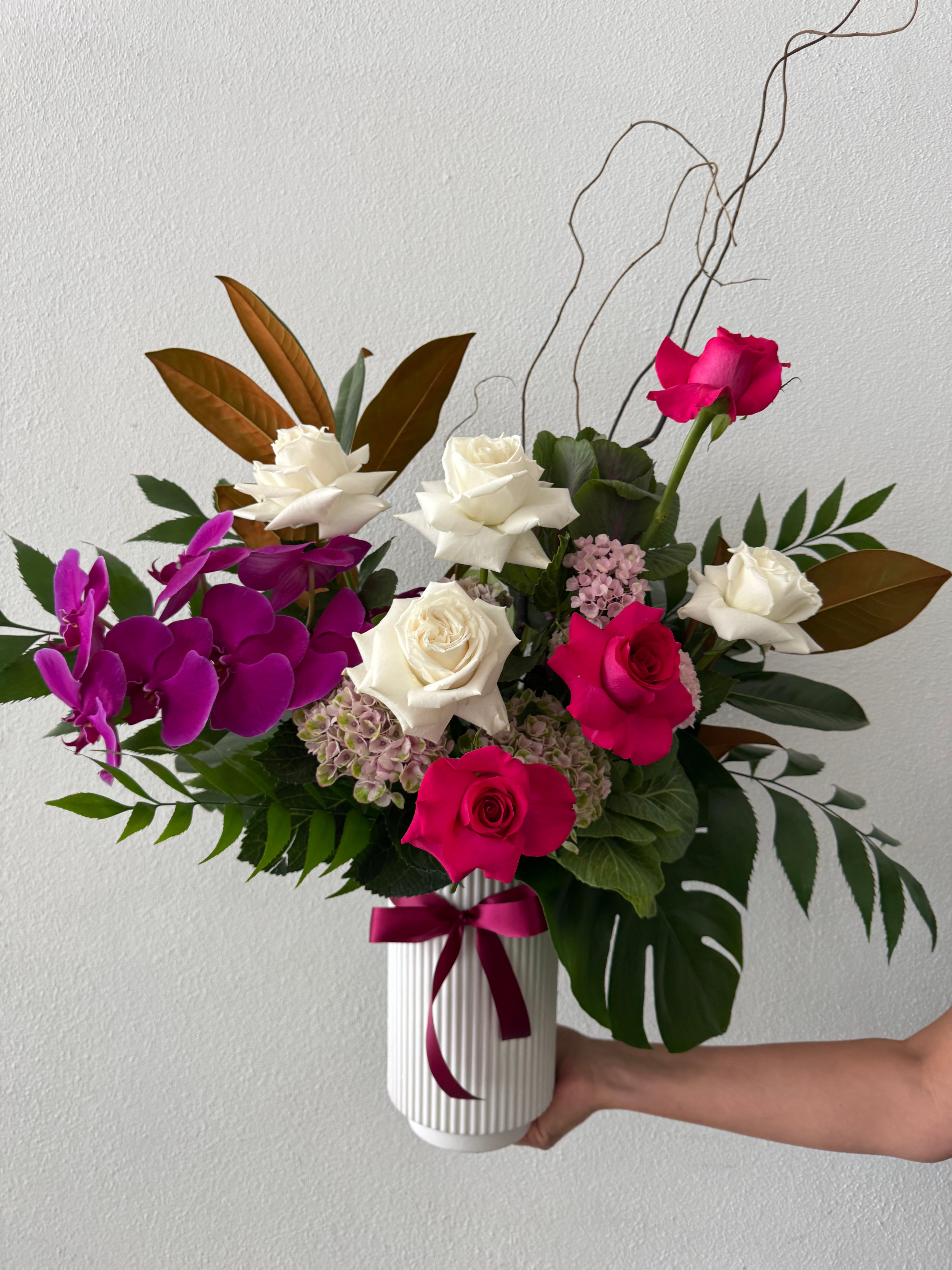 A floral arrangement featuring roses in shades of white and red roses with pink and magenta flower and green foliage, held in a white vase.