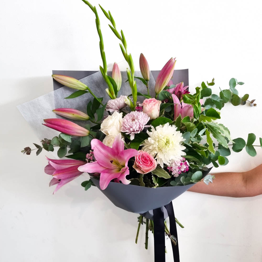 A hand-held bouquet featuring soft pastel mixed seasonal blooms, with a variety of flowers including pink lilies and white and pink roses, with green foliage